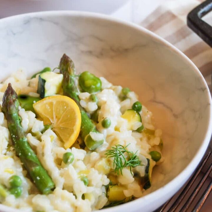 two bowls of risotto with asparagus, broad beans, peas, fresh dill and a slice of lemon. A silver fork sits beside the bowl at the front.