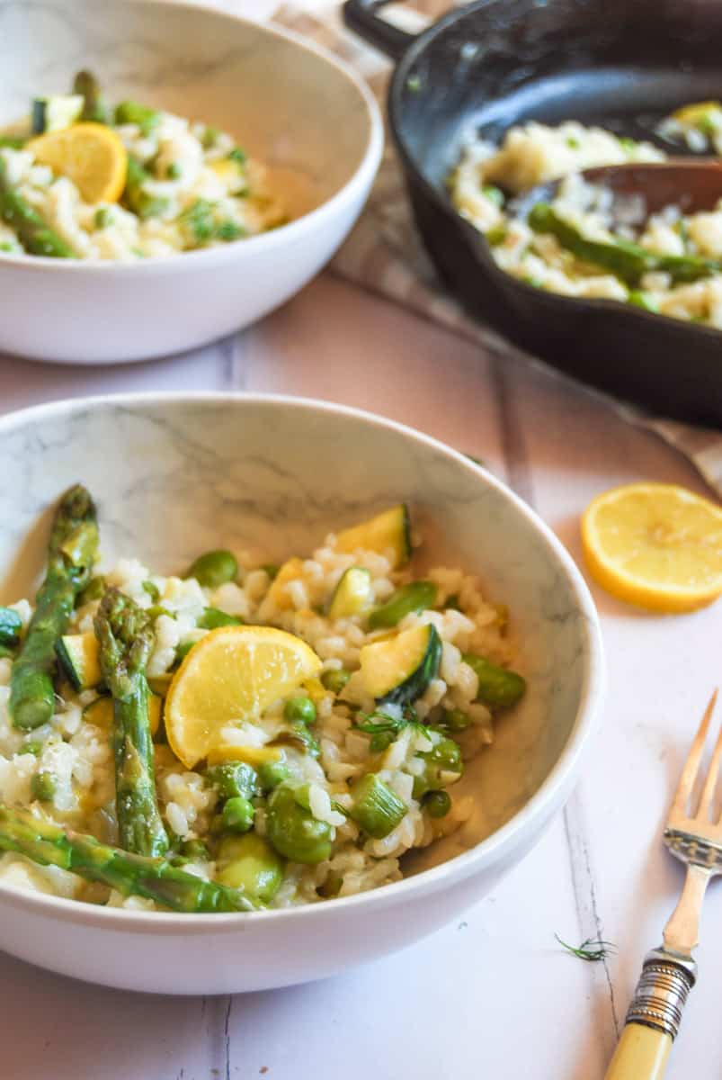 two white bowls of risotto with asparagus, broad beans, diced courgettes and peas with a slice of lemon, a gold handled silver fork. A black pan of risotto can be partially seen in the background.