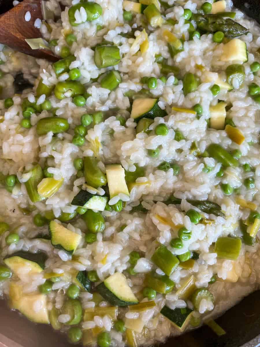 A pan of risotto with broad beans, diced courgette, sliced leeks and peas with a wooden spoon.