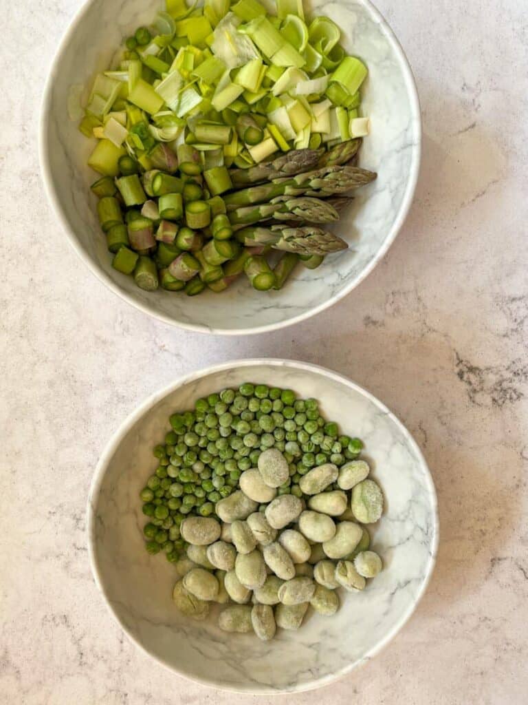 two white bowls of frozen broad beans, peas, chopped asparagus spears and sliced leek.