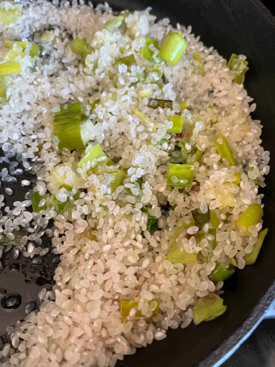 Risotto rice and sliced leeks in a black saucepan.