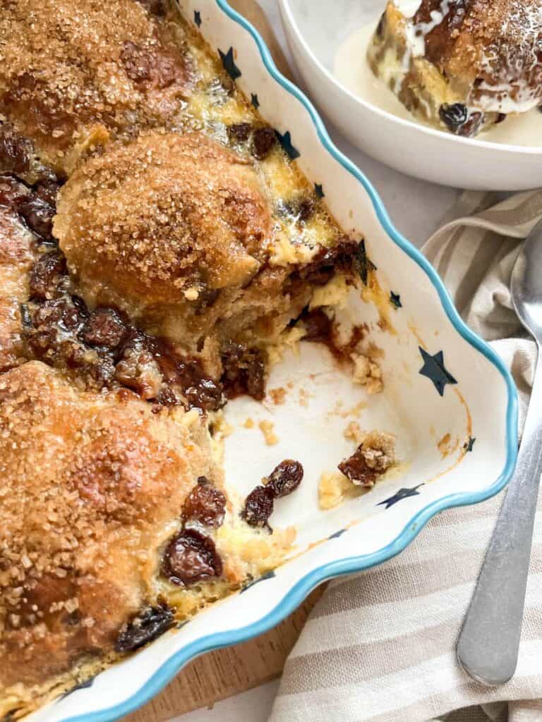 a blue and white star dish of hot cross bun bread pudding with raisins and chocolate chips, a beige and white striped tea towel and a silver spoon.