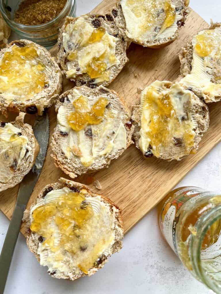 hot cross buns split open and spread with butter and marmalade on a wooden chopping board.