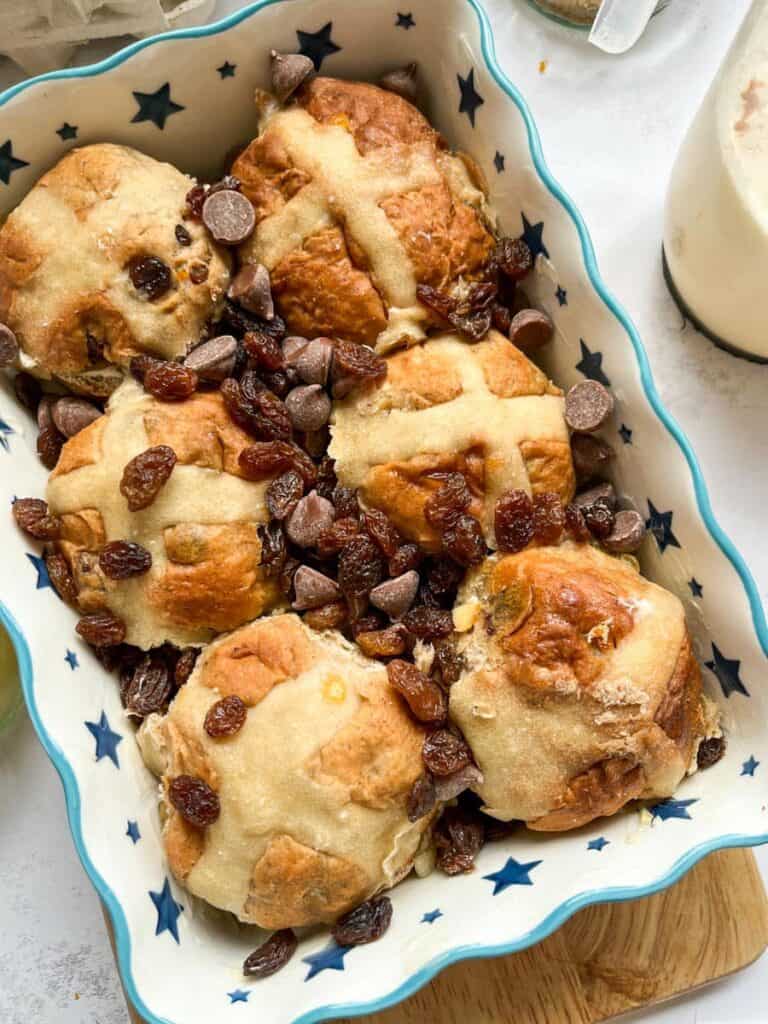 six hot cross buns scattered with chocolate chips and raisins in a blue and white star patterned dish on a wooden chopping board.