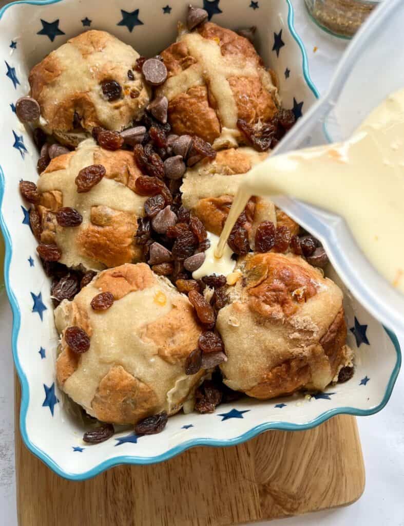 a jug of beaten eggs, milk and cream being poured into a blue and white star patterned dish of hot cross buns, raisins and chocolate chips.