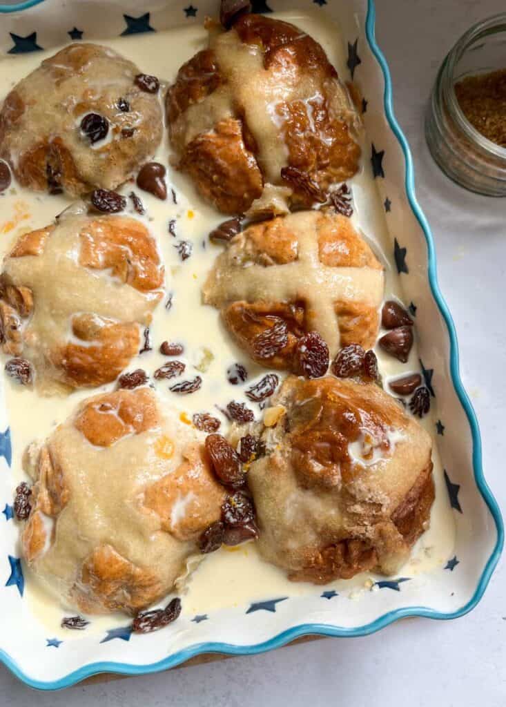 six hot cross buns in a blue and white star patterned dish scattered with raisins and chocolate chips soaking in a custard mixture.