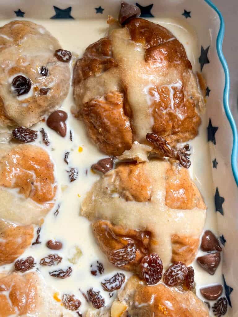 six hot cross buns soaked in custard and scattered with chocolate chips and raisins in a blue and white star patterned dish.