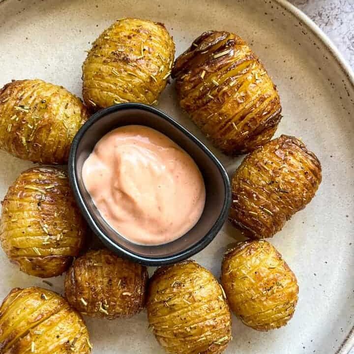 A plate of hasselback potatoes sprinkled with dried rosemary on a beige plate and a black pot of Sriracha mayonnaise.