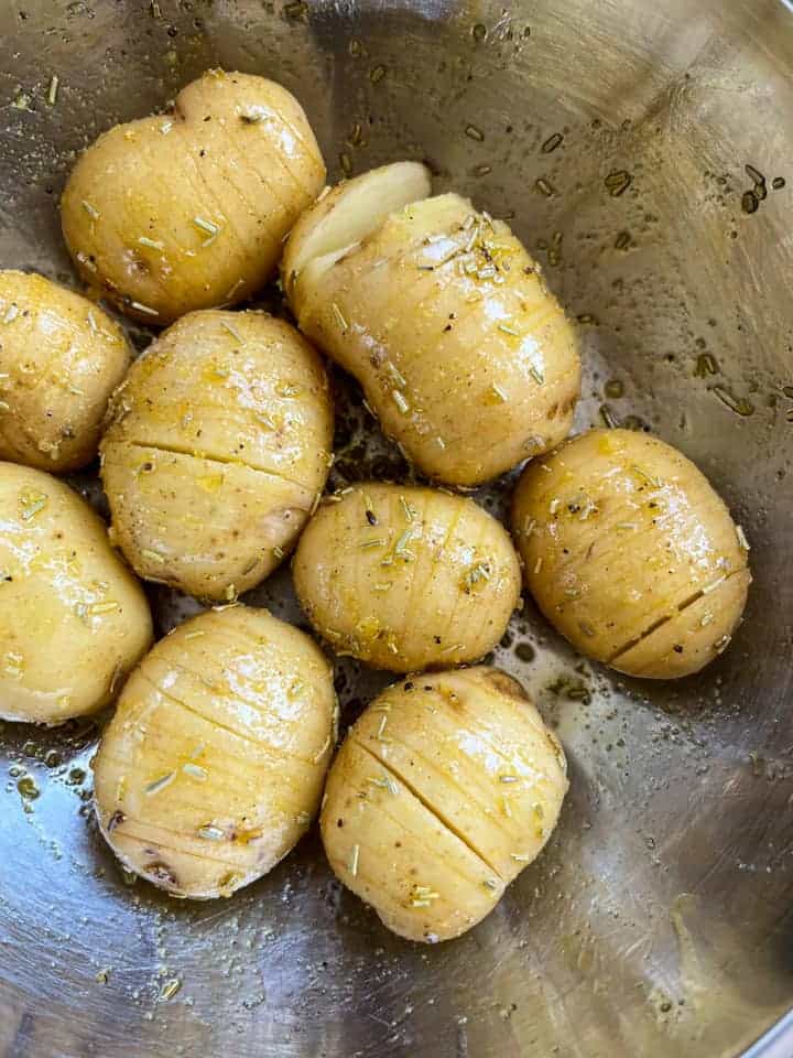 nine sliced potatoes coated with oil, salt and pepper and dried rosemary in a silver bowl.