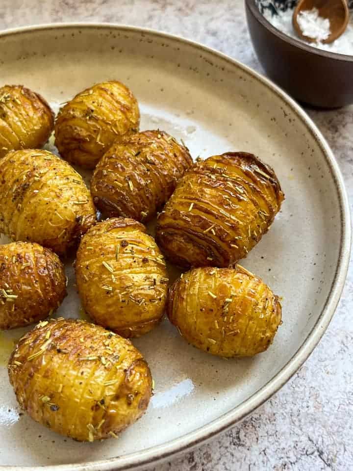 Hasselback potatoes sprinkled with dried rosemary on a beige plate and a wooden pot of flaked salt.