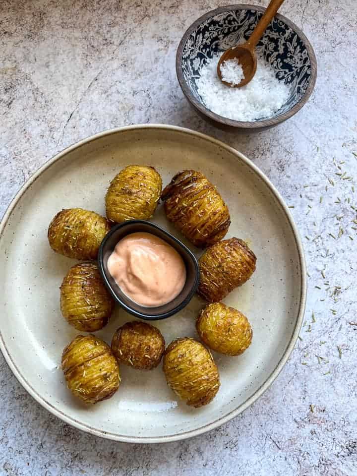 nine hasselback potatoes on a beige plate with a black pot of Sriracha mayonnaise and a wooden pot of flaked sea salt with a small wooden spoon.