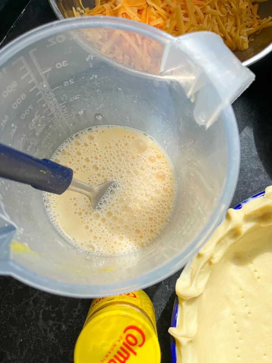 a plastic jug of beaten eggs with a blue fork, a silver bowl of grated cheese, a yellow jar of mustard and a blue and white pie dish lined with pastry.