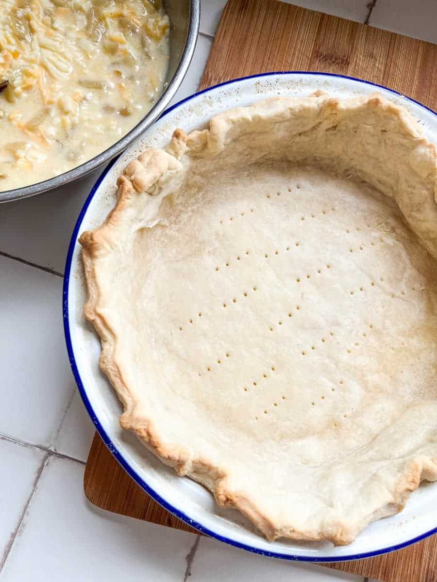 a blue and white metal pie dish lined with a pastry case on a wooden serving board and a bowl of grated cheese, eggs and milk.