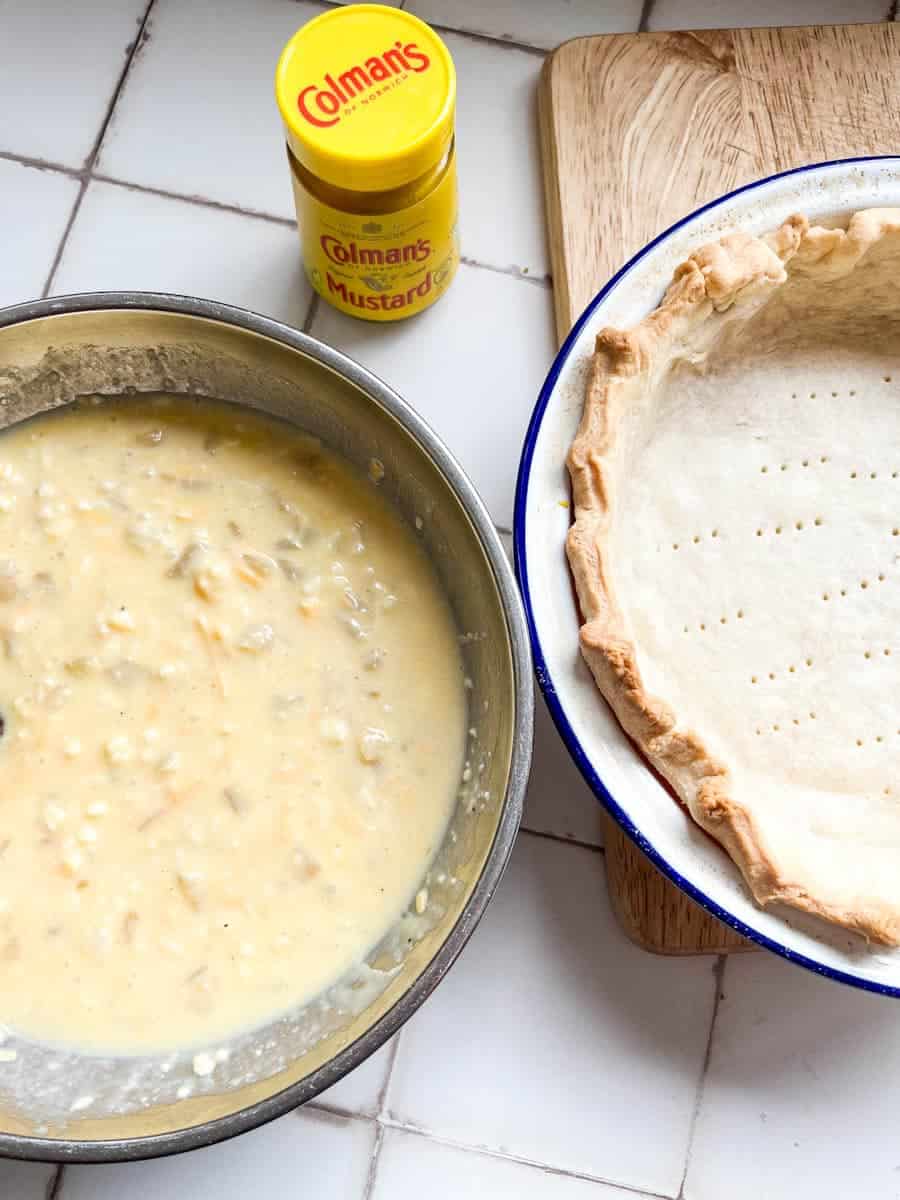 a silver bowl of grated cheese, eggs, milk and diced onion, a blue and white pie dish lined with a shortcrust pastry dish and a yellow jar of mustard.