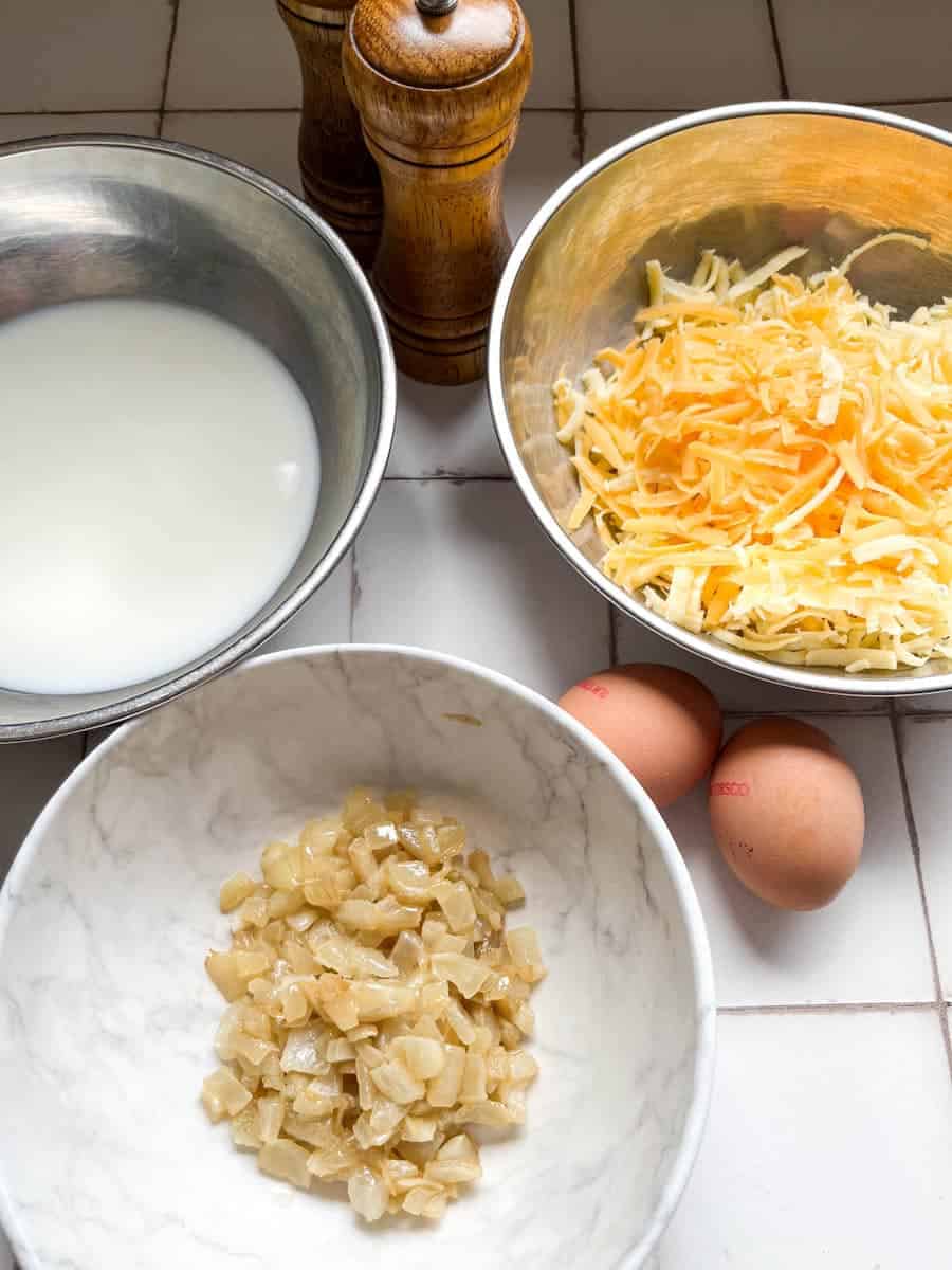 a white marble bowl of cooked diced onions, a silver bowl of grated cheese, a silver bowl of milk, two eggs and wooden salt and pepper mills.