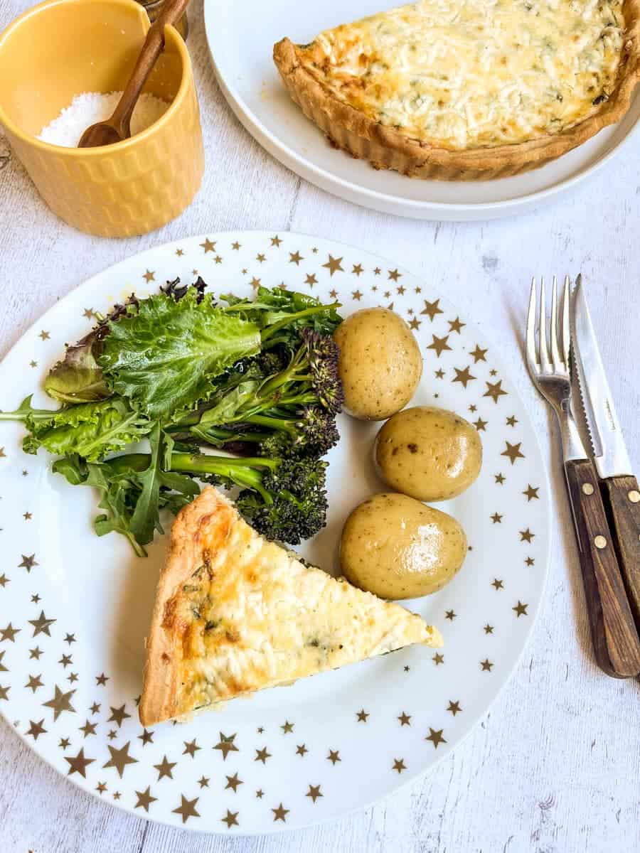 a slice of quiche, boiled new potatoes, broccoli and green salad leaves on a white and gold star plate, a knife and fork with wooden handles, a yellow pot of sea salt with a wooden spoon and a quiche sitting on a large white plate.