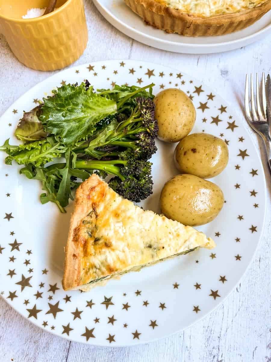 a slice of quiche, boiled new potatoes, broccoli and green salad leaves on a white and gold star plate.