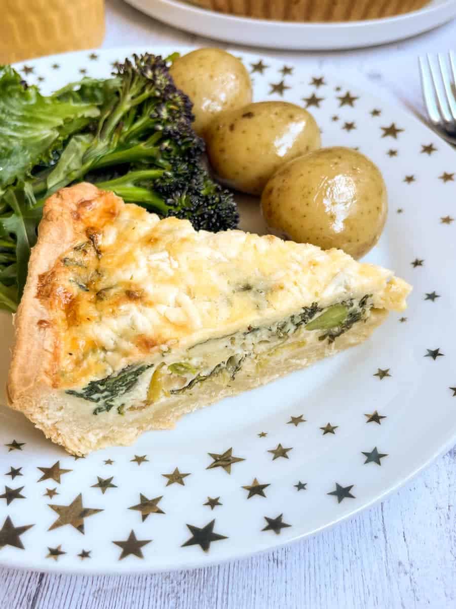 a slice of quiche with leeks and spinach, boiled new potatoes and broccoli on a white and gold star plate.