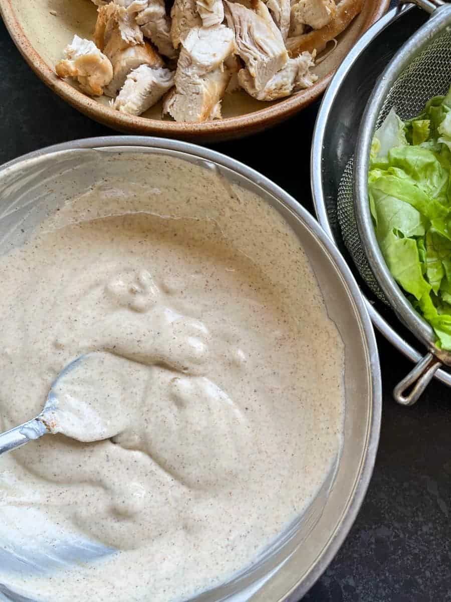 a silver bowl of mayonnaise, yoghurt and curry powder stirred together with a silver spoon, a bowl of cooked chopped chicken and chopped lettuce in a sieve.