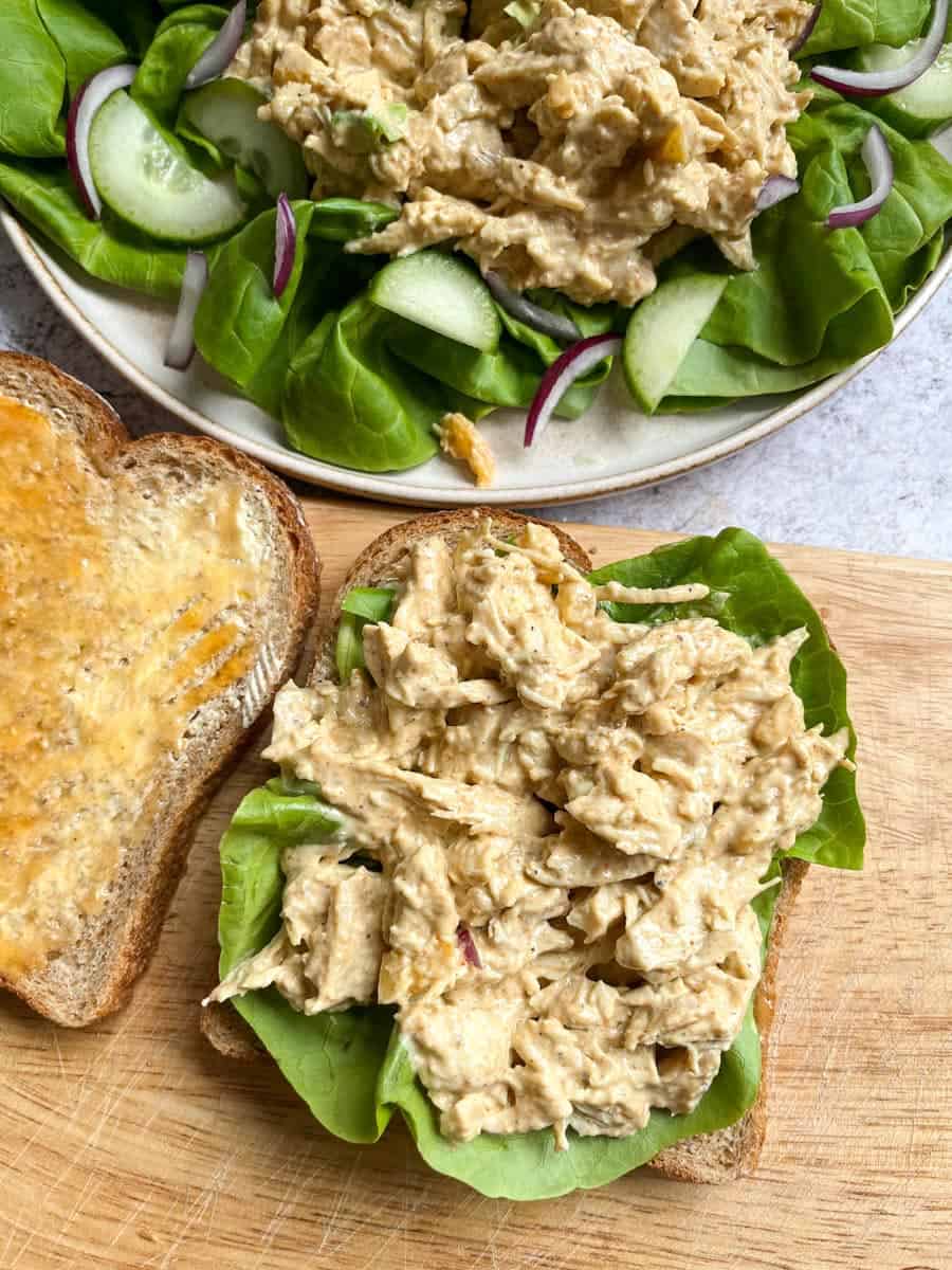 two slices of brown bread on a chopping board topped with lettuce and cold chicken in a curry sauce. A plate of chicken salad with lettuce, cucumbers and red onions sits above the bread.