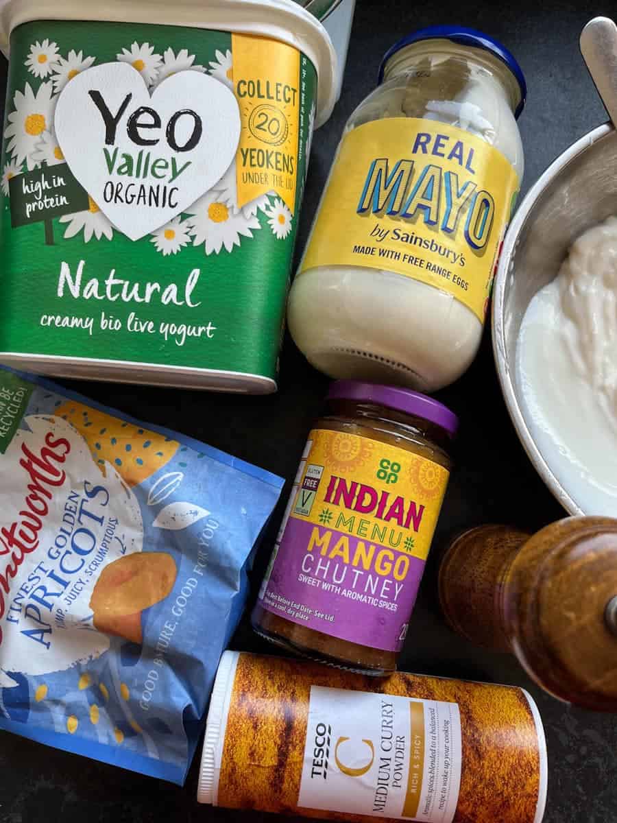 A tub of natural yoghurt, a jar of mayonnaise, a jar of mango chutney, a packet of dried apricots, a tub of curry powder, a wooden pepper mill and a silver bowl of yoghurt.