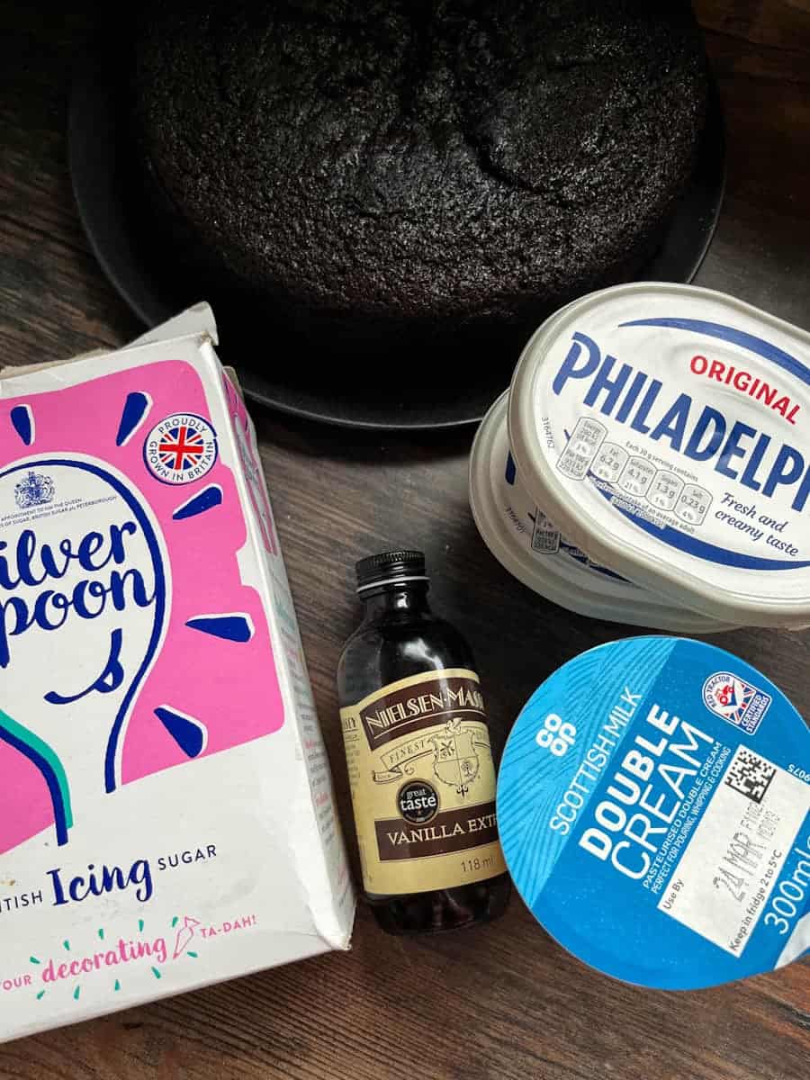 A chocolate cake on a black plate, two tubs of Philadelphia cheese, a box of icing sugar, a bottle of vanilla extract and a pot of double cream.