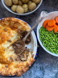 A minced beef pie with a flaky pastry crust in a round pie dish with a silver spoon, a bowl of peas and sliced carrots and a bowl of potatoes.