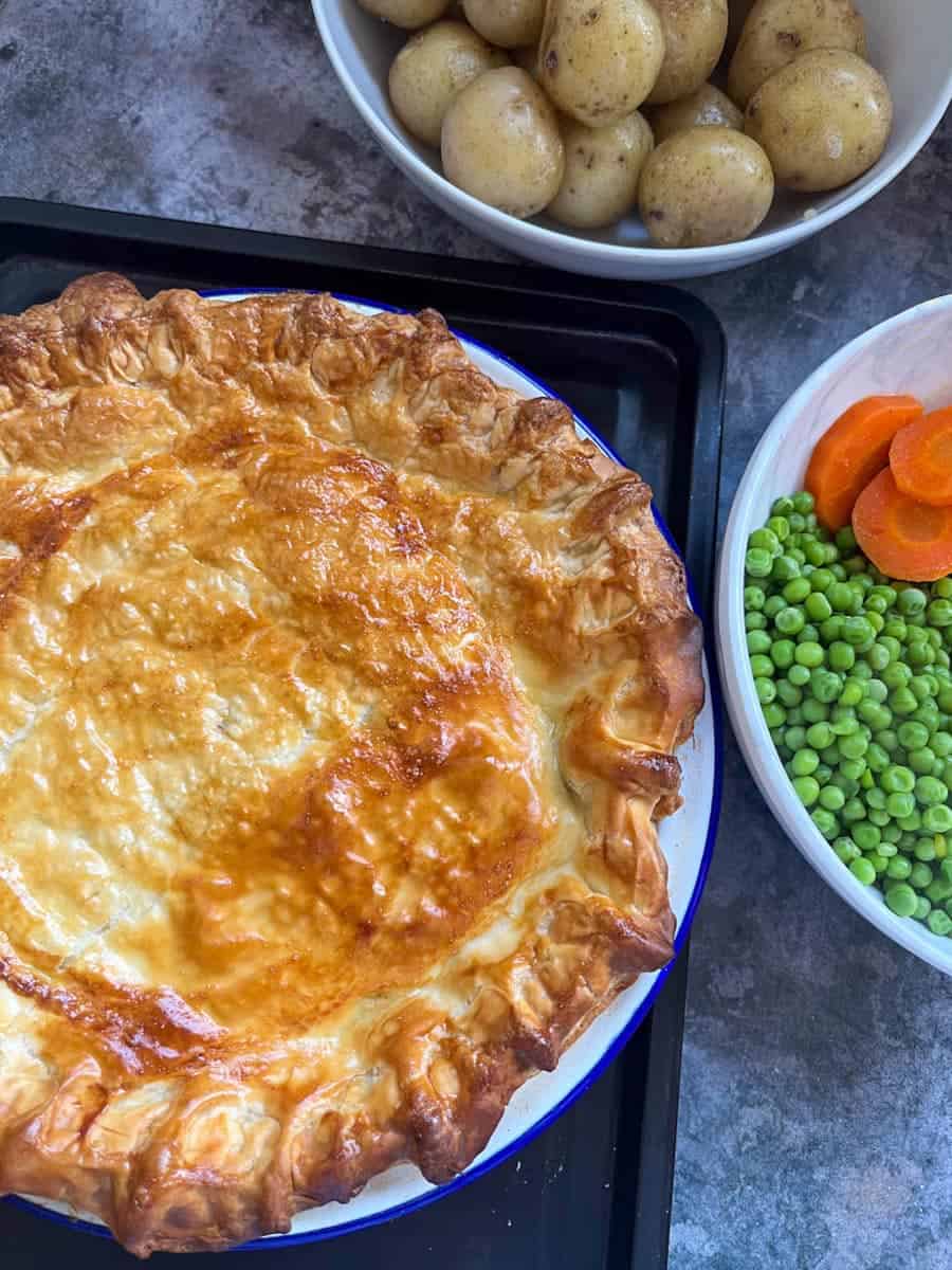 a mince beef pie with a puff pastry lid, a white bowl of peas and carrots and a bowl of new potatoes.