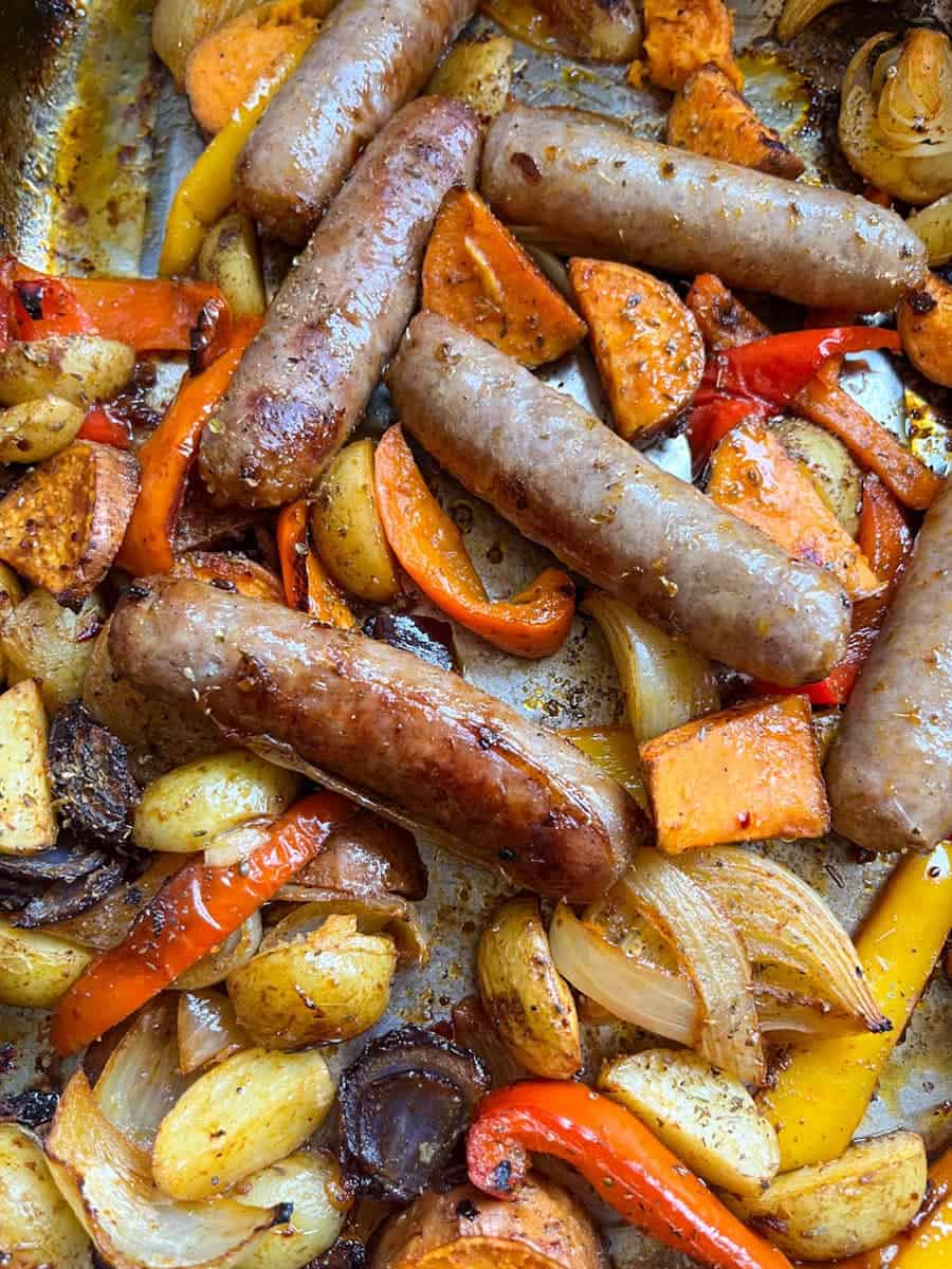 a pan of cooked pork sausages, sweet potatoes, sliced red and yellow peppers and sliced red and white onion.