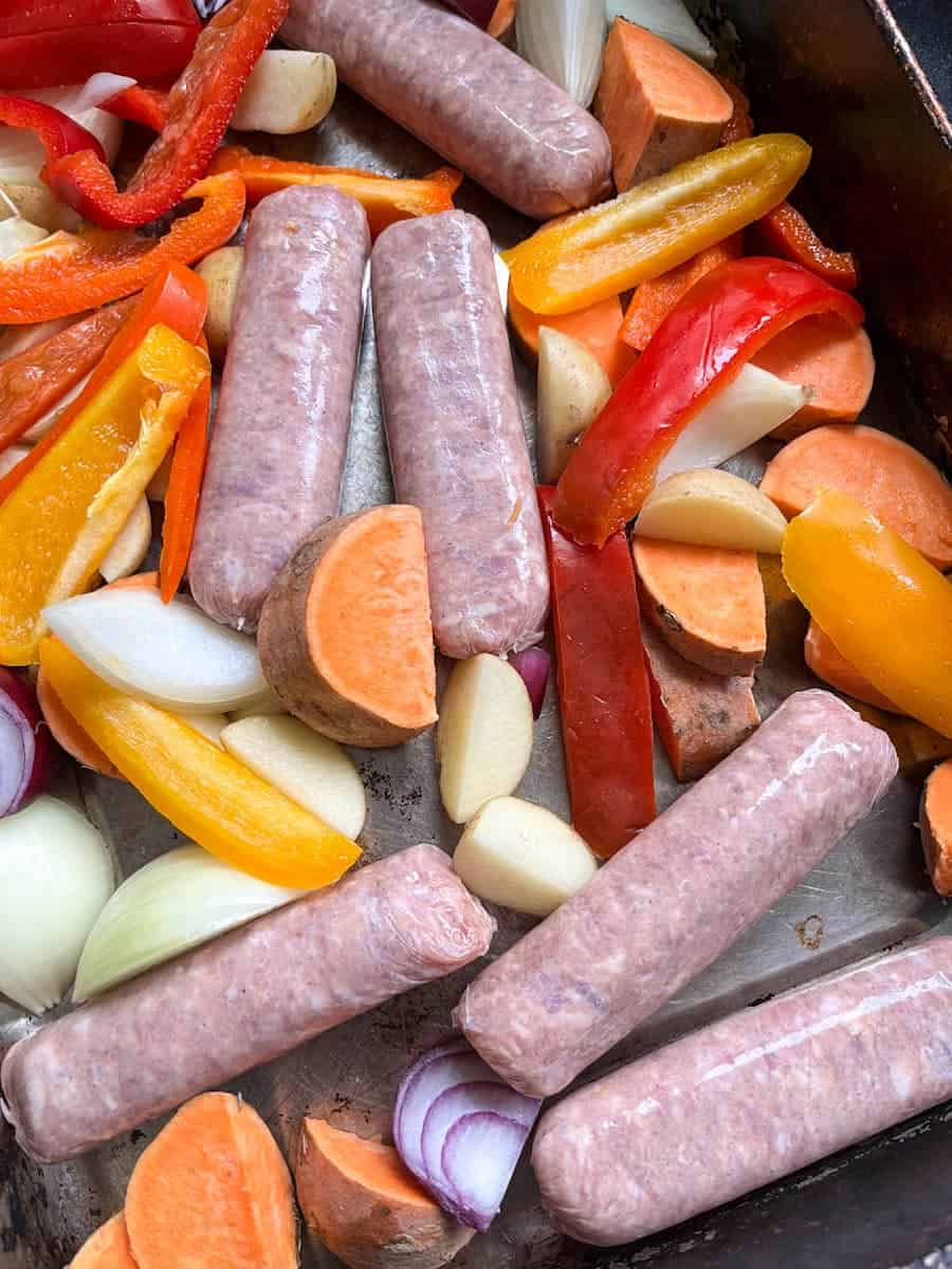 a tray of uncooked pork sausages, sweet potatoes, sliced red and yellow peppers and sliced onion.