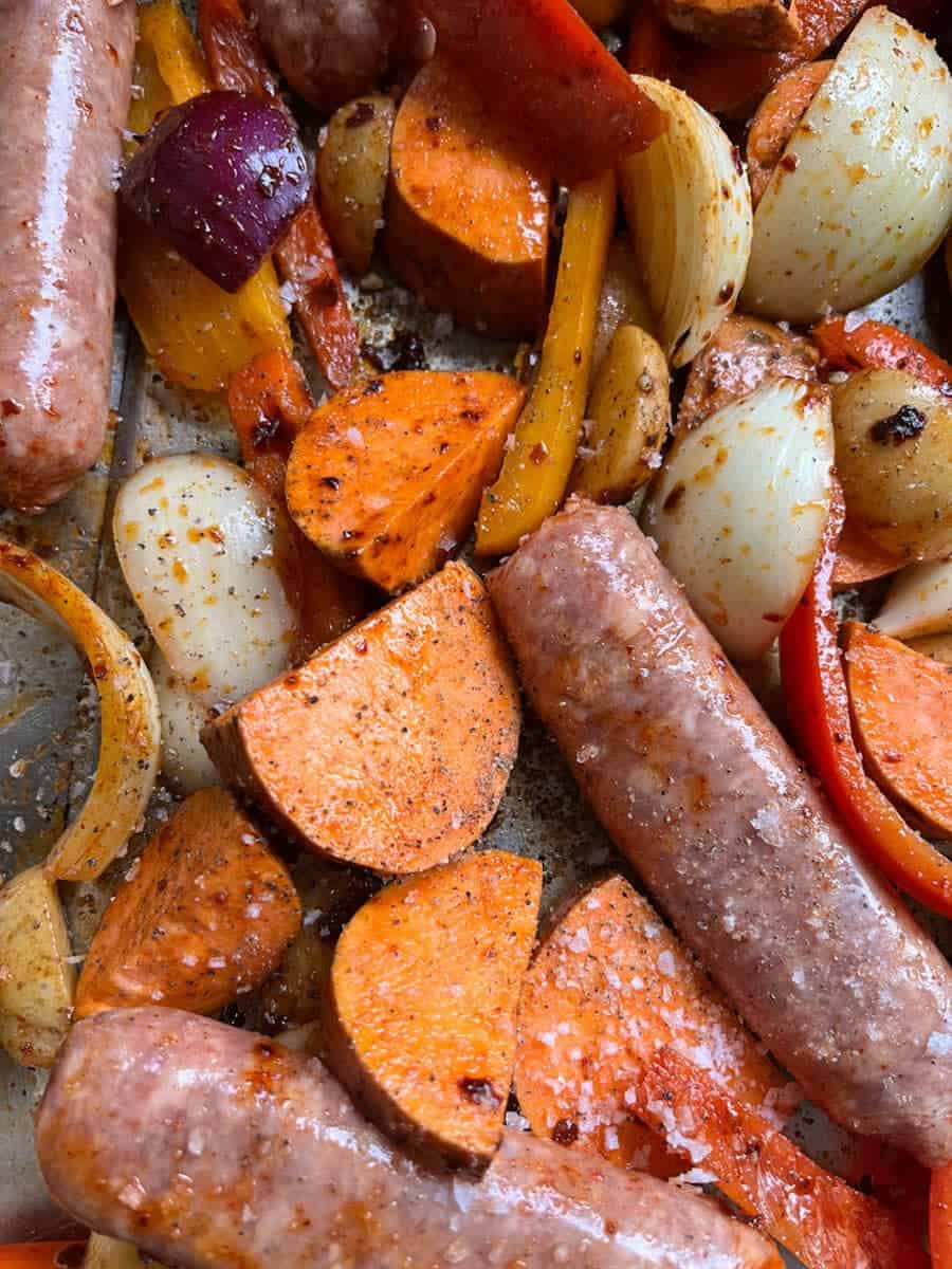 a close up picture of a tray of uncooked sausages, sweet potatoes, red onion and sliced red and yellow peppers.