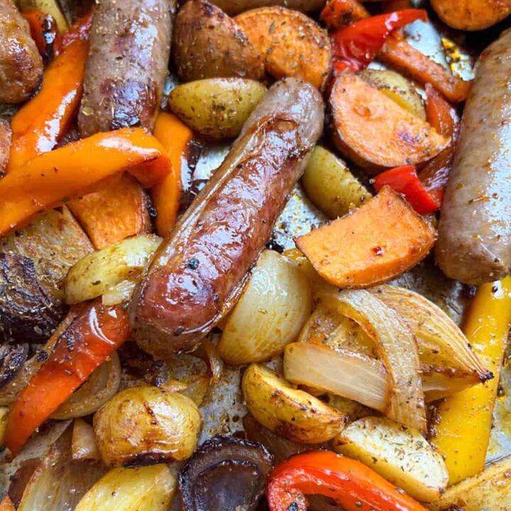 a roasting tin with cooked pork sausages, sliced red and white onion, sliced red and yellow peppers and potato wedges.