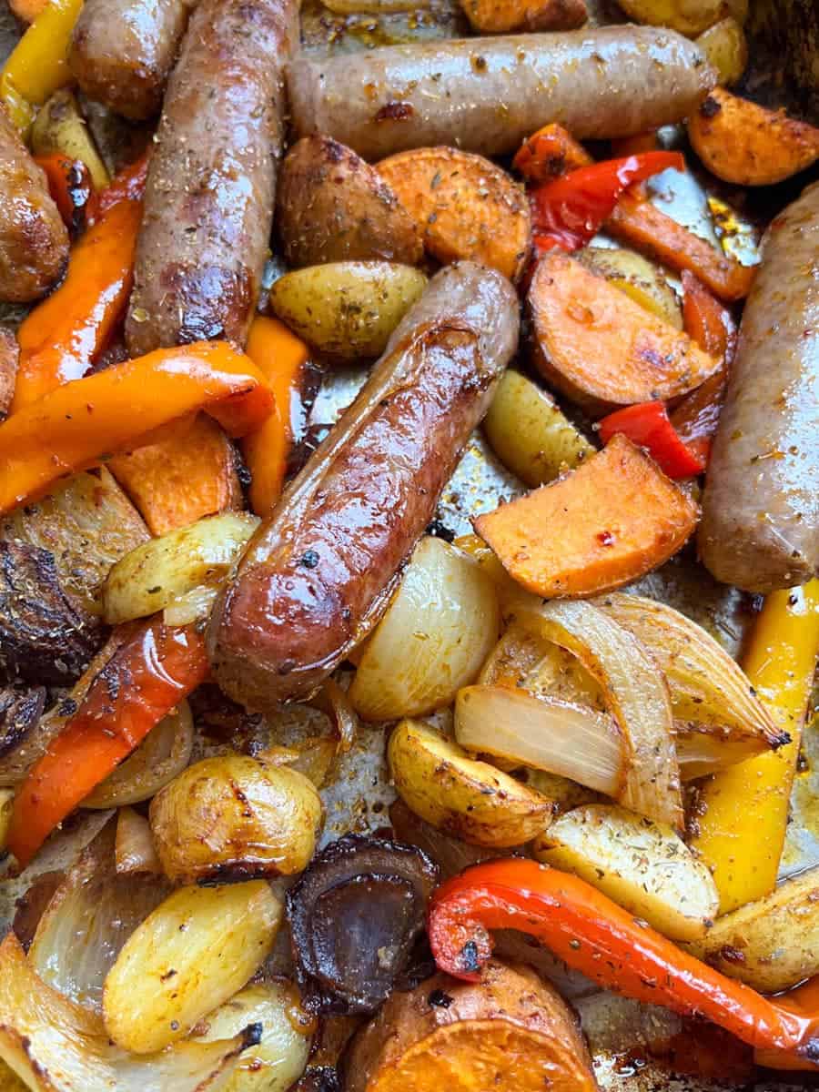 a roasting tin with cooked pork sausages, sliced red and white onion, sliced red and yellow peppers and potato wedges.