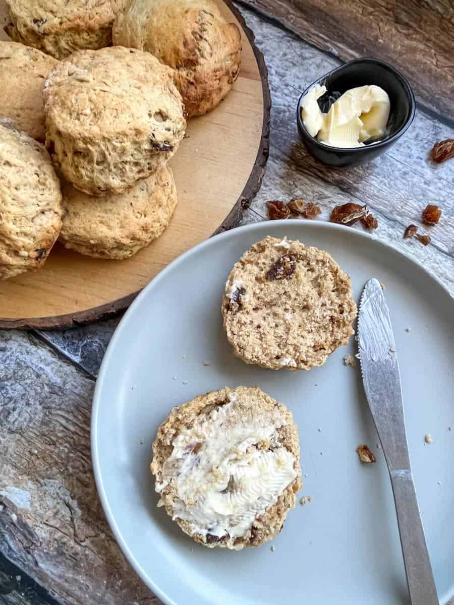 a date and walnut scone split in half and spread with butter on a grey plate with a knife, a small black dish of butter and a batch of scones on a round wooden serving board.
