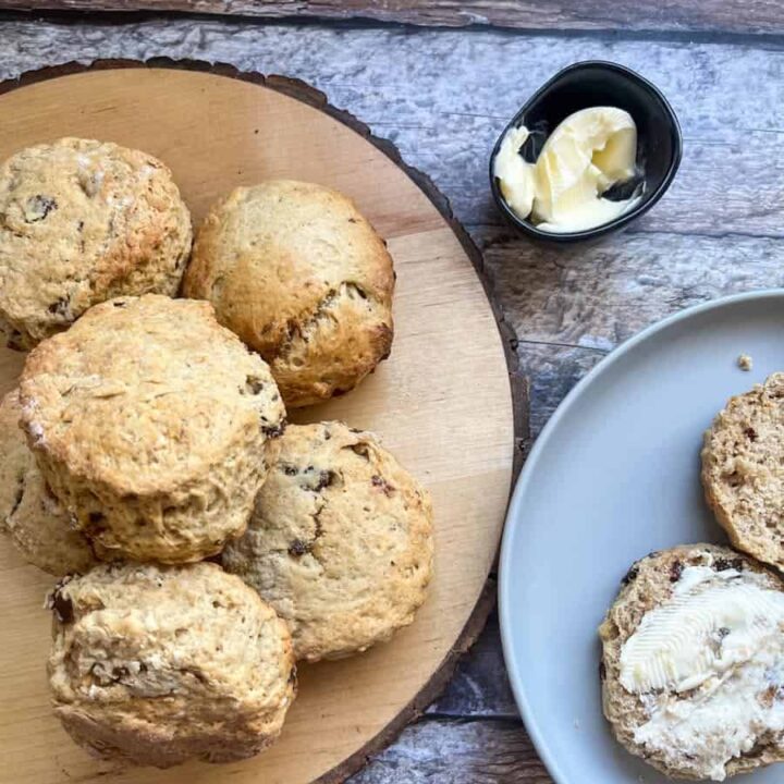 six date and walnut scones on a round wooden serving board, a scone split in half and spread with butter on a grey plate and a small black dish of butter on a wooden backdrop.