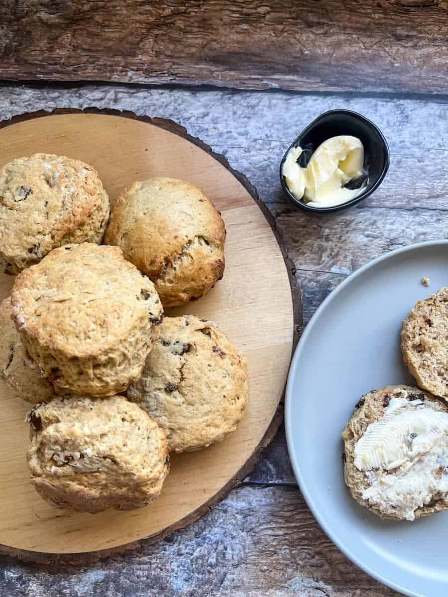 six date and walnut scones on a round wooden serving board, a scone split in half and spread with butter on a grey plate and a small black dish of butter on a wooden backdrop.