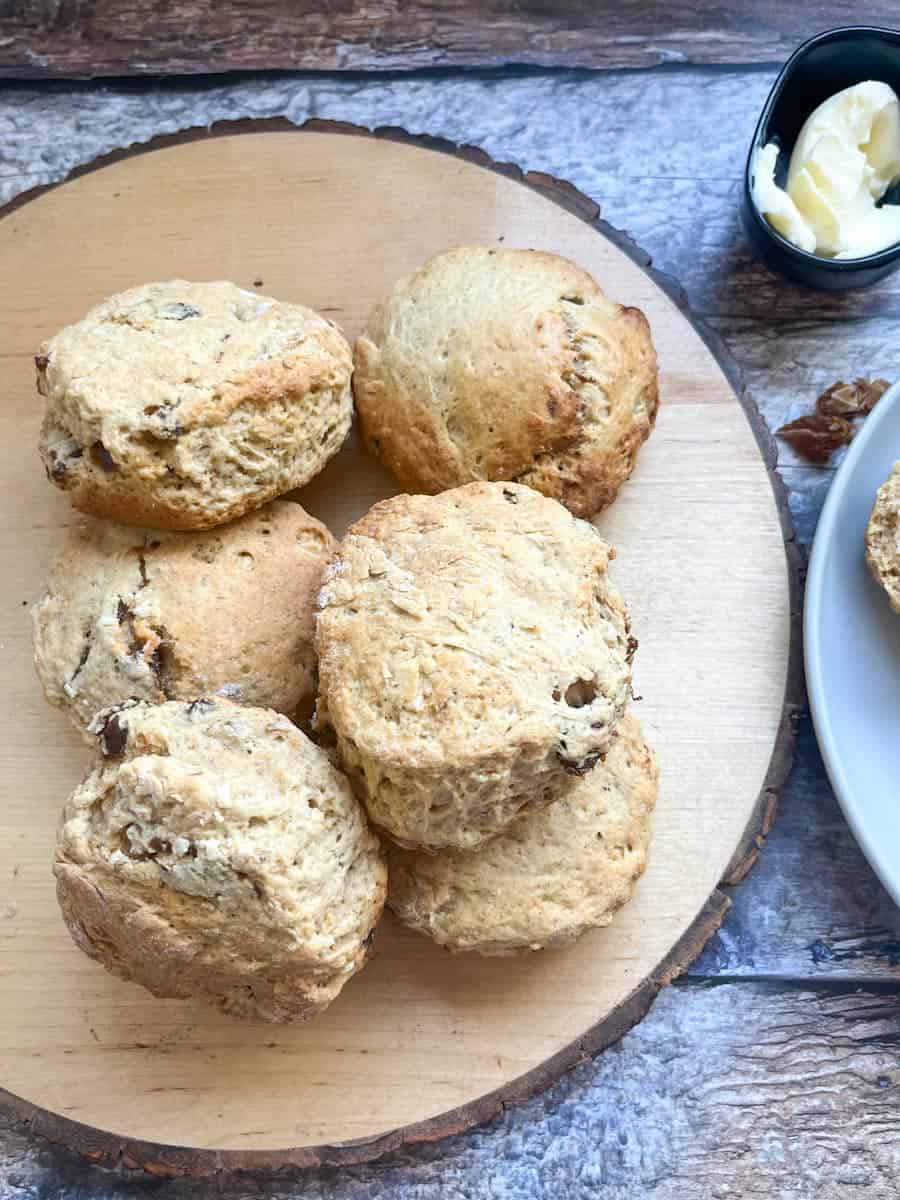 six scones with dates and walnut on a round wooden serving board and a small black dish of butter on a wooden backdrop.