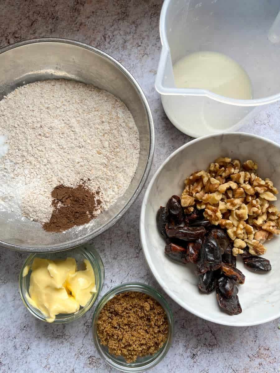 a white bowl of dates and walnuts, a silver bowl of flour and mixed spice, a jug of milk, a small dish of butter and a dish of brown sugar.