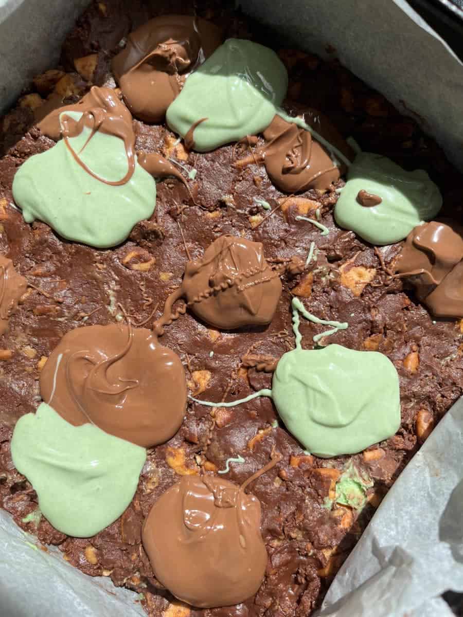 melted milk and green coloured chocolate dropped in rough circles onto a traybake made from crushed biscuits and chocolate.