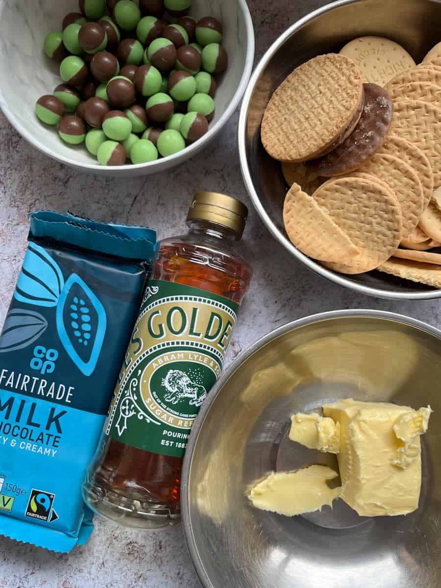a silver bowl of butter, a bowl of biscuits, a white bowl of green and white chocolate balls, a bottle of golden syrup and a bar of milk chocolate.