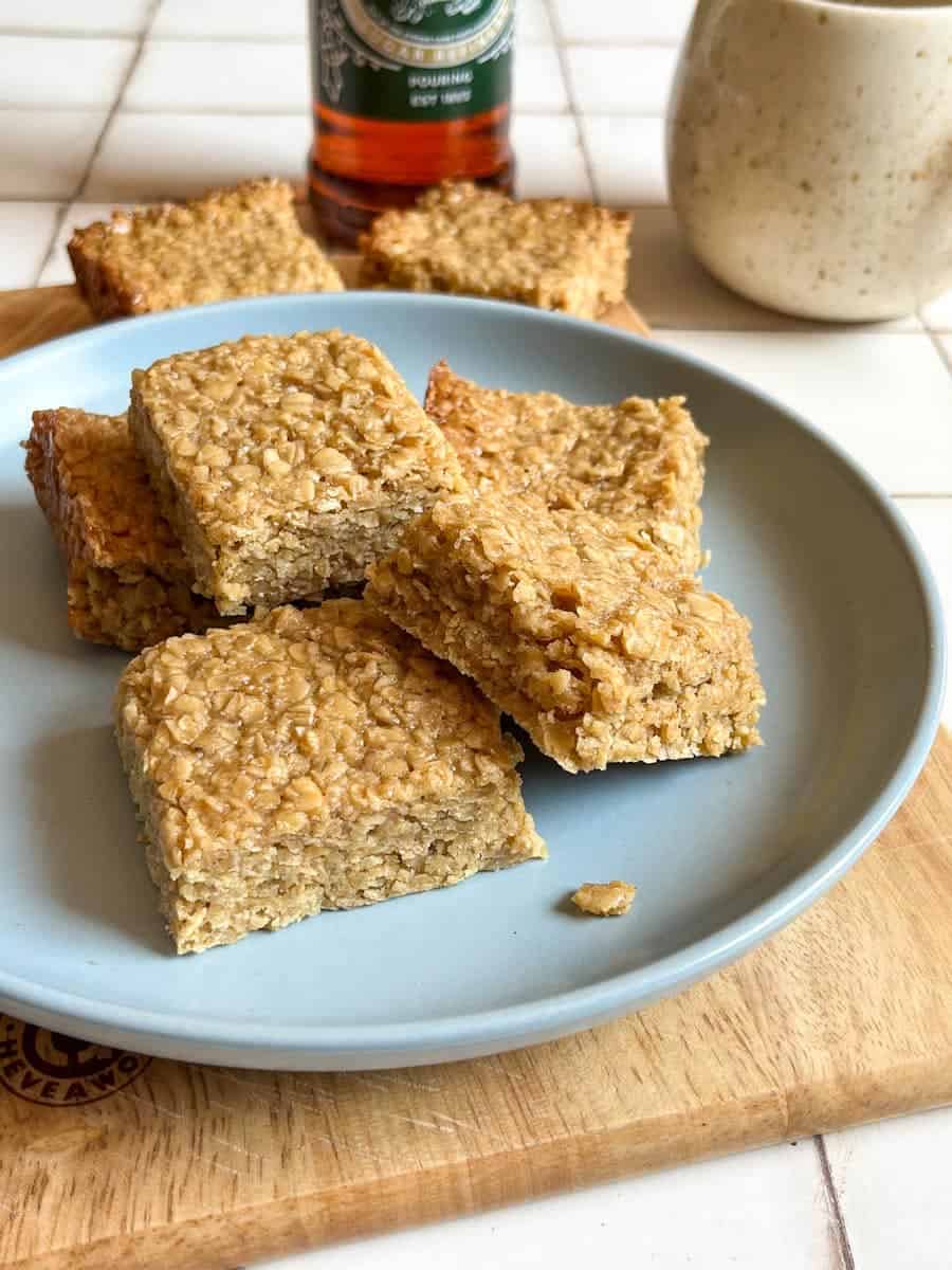 a plate of oat flapjack bars on a blue plate placed on a wooden chopping board. A beige mug and a bottle of golden syrup are in the background.