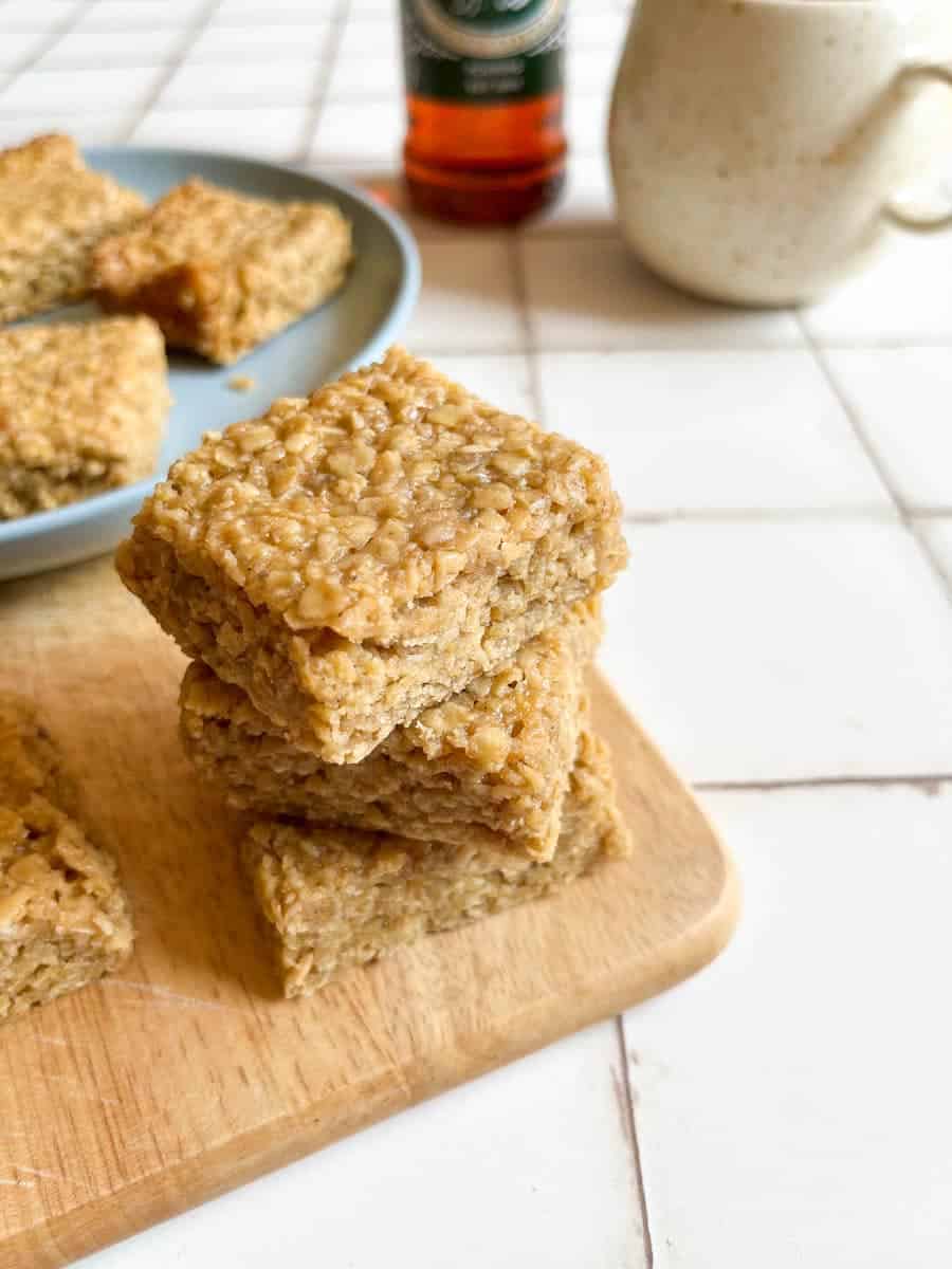 three flapjack oat bars stacked on top of each other on a wooden board. A blue plate with more oat bars can be seen in the background along with a beige mug and a bottle of golden syrup.