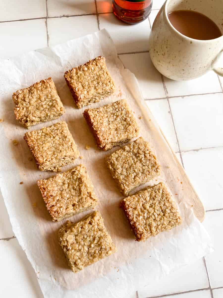 eight flapjacks on a piece of baking paper and a large beige mug of tea on a white tiled surface.