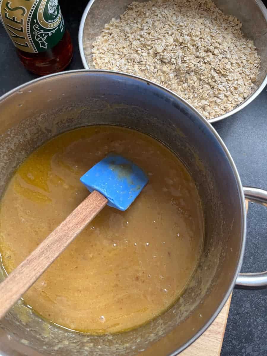 A silver pan of melted butter, sugar and syrup with a blue wooden handled spatula, a silver bowl of oats and a bottle of golden syrup.