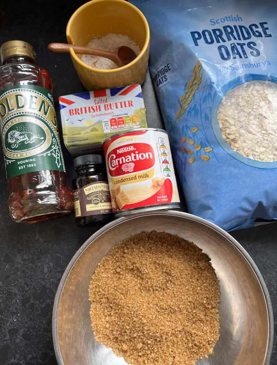 a silver bowl of brown sugar, a blue packet of rolled oats, a tin of condensed milk, a jar of vanilla extract, a bottle of golden syrup, a block of butter and a yellow jar of salt with a small wooden spoon.