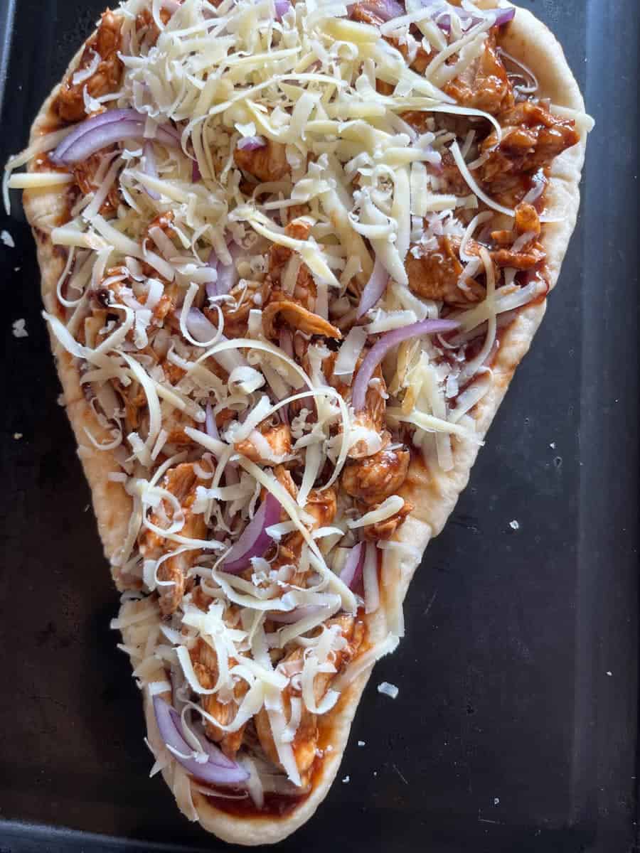 an uncooked Naan bread pizza with chicken, bbq sauce, grated cheese and sliced red onion on a baking tray.