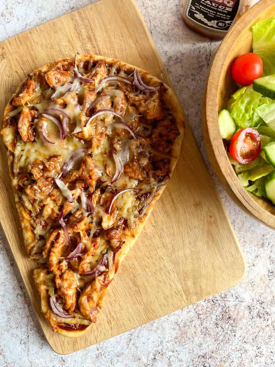 a Naan bread pizza with chicken, bbq sauce, sliced red onion and melted cheese on a wooden chopping board and a wooden salad bowl of shredded lettuce, cucumber and tomatoes.
