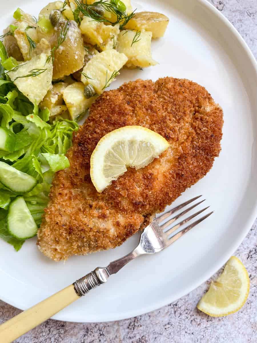 a chicken fillet covered in golden breadcrumbs topped with a lemon slice on a white plate with lettuce, cucumber and potato salad.