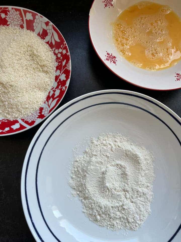 a white bowl of flour, a red and white bowl of beaten egg and a red and a white bowl of panko breadcrumbs.