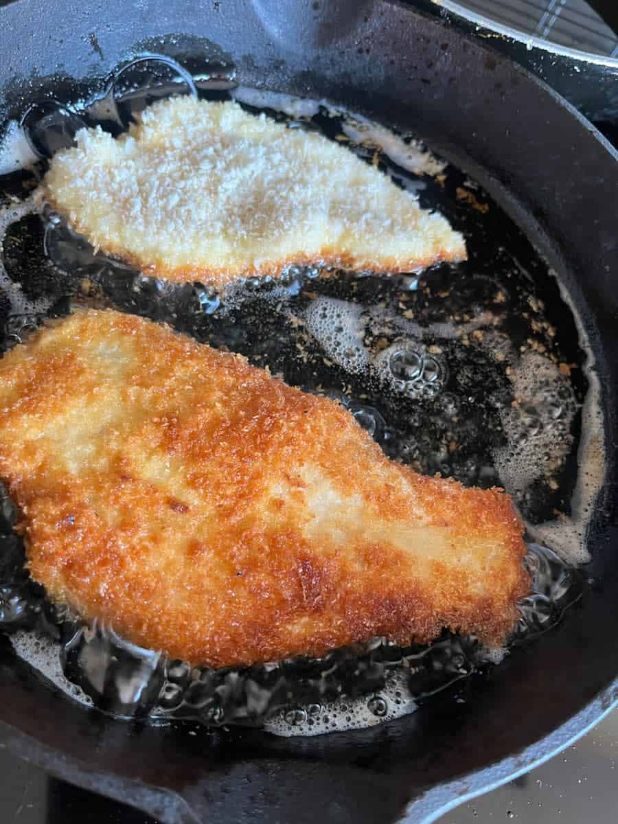 two chicken fillets covereed in breadcrumbs cooking in a black saucepan.
